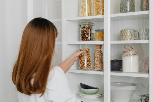 Woman organizing pantry by placing dry goods into labeled glass jars on a white minimalist shelf with mugs, bowls, and ceramic containers – Home Life Manila